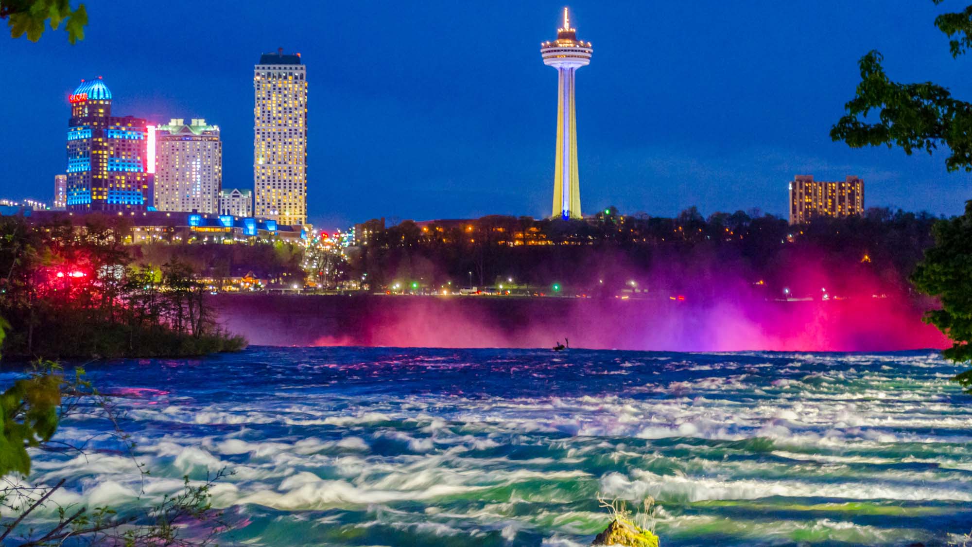 Niagara Falls oplyst af farvestrålende lys og med Skylon Tower i baggrunden.