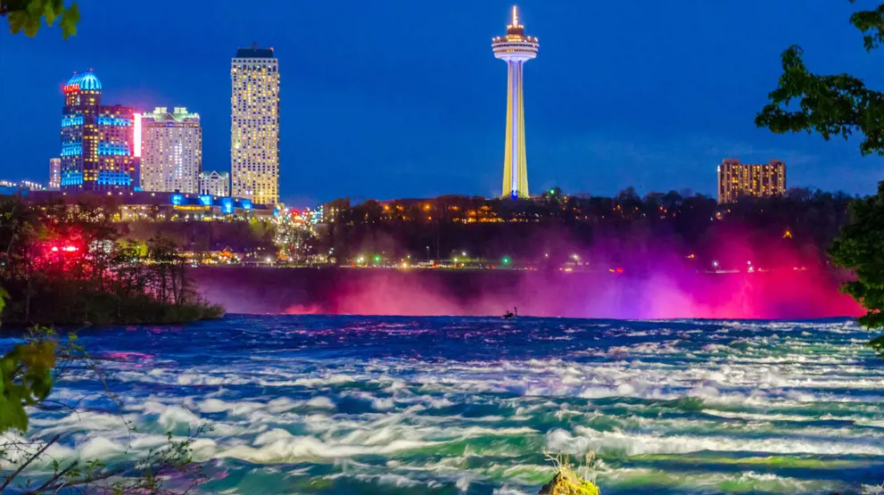 Niagara Falls oplyst af farvestrålende lys og med Skylon Tower i baggrunden.